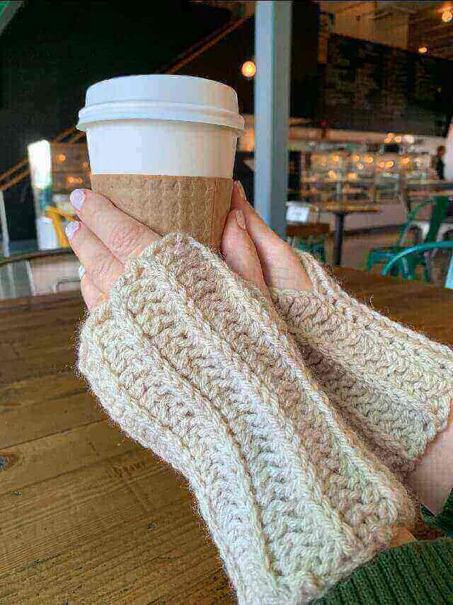 A pair of hands wearing crocheted finger-less mitts holding a takeout coffee cup.