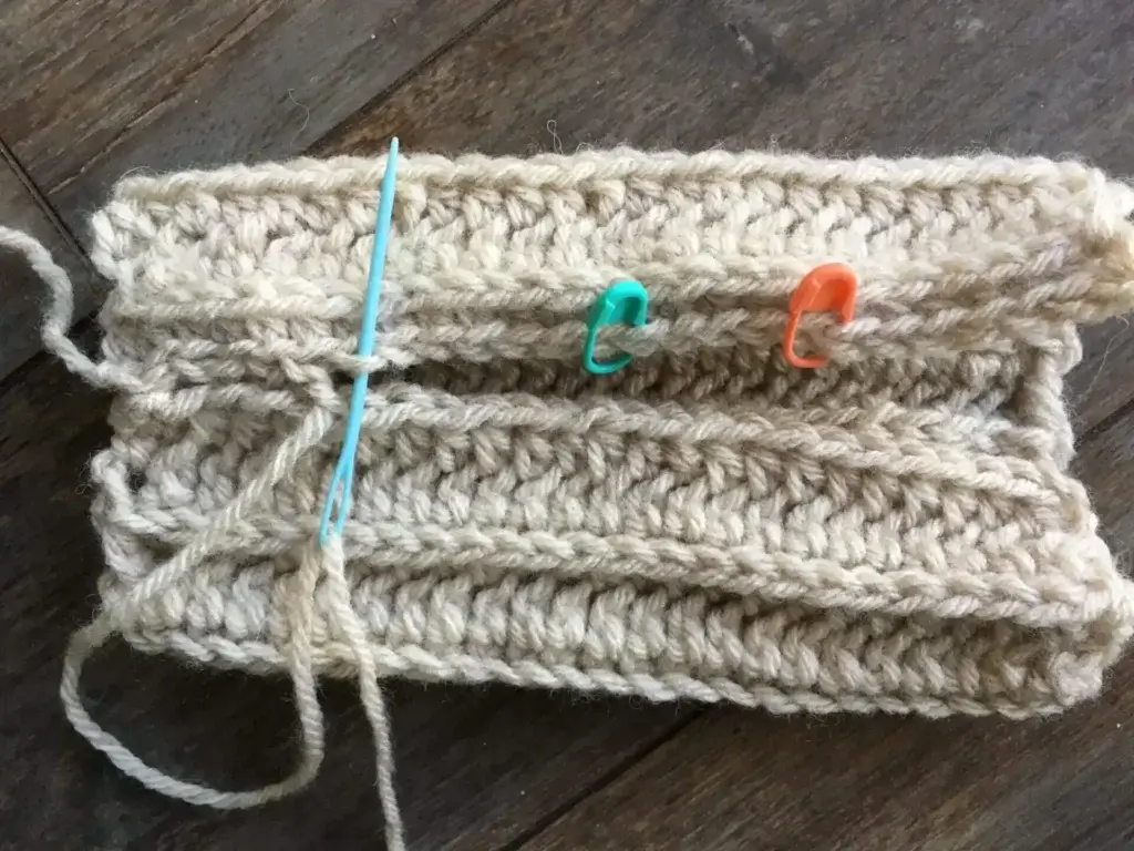 A rectangular piece of crocheted fabric with orange and blue stitch markers attached. The stitch markers indicate where the stitches will be left open to create a thumb opening for fingerless mitts.