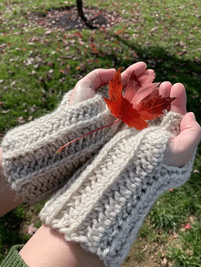 A pair of hands wearing crocheted finger-less mitts holds a red maple leaf.