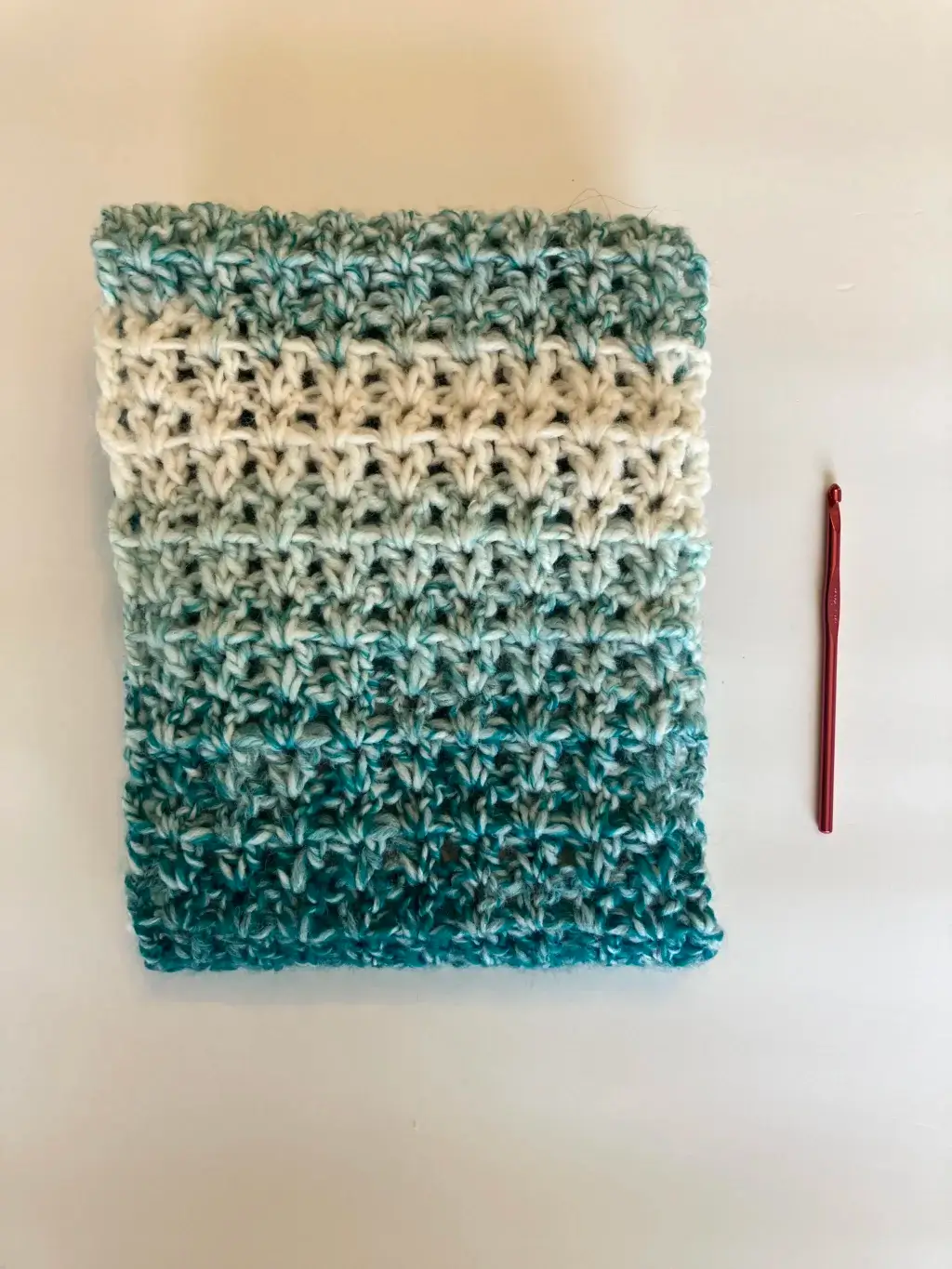 A crochet infinity scarf is folded and sitting on a white background. A red crochet hook is next to it.